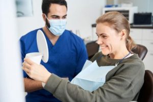 Dental professional in blue scrubs and mask assisting a smiling patient in a dental chair, holding a mirror to view her teeth, emphasizing patient comfort and care at Farber Center for Periodontics & Dental Implants.